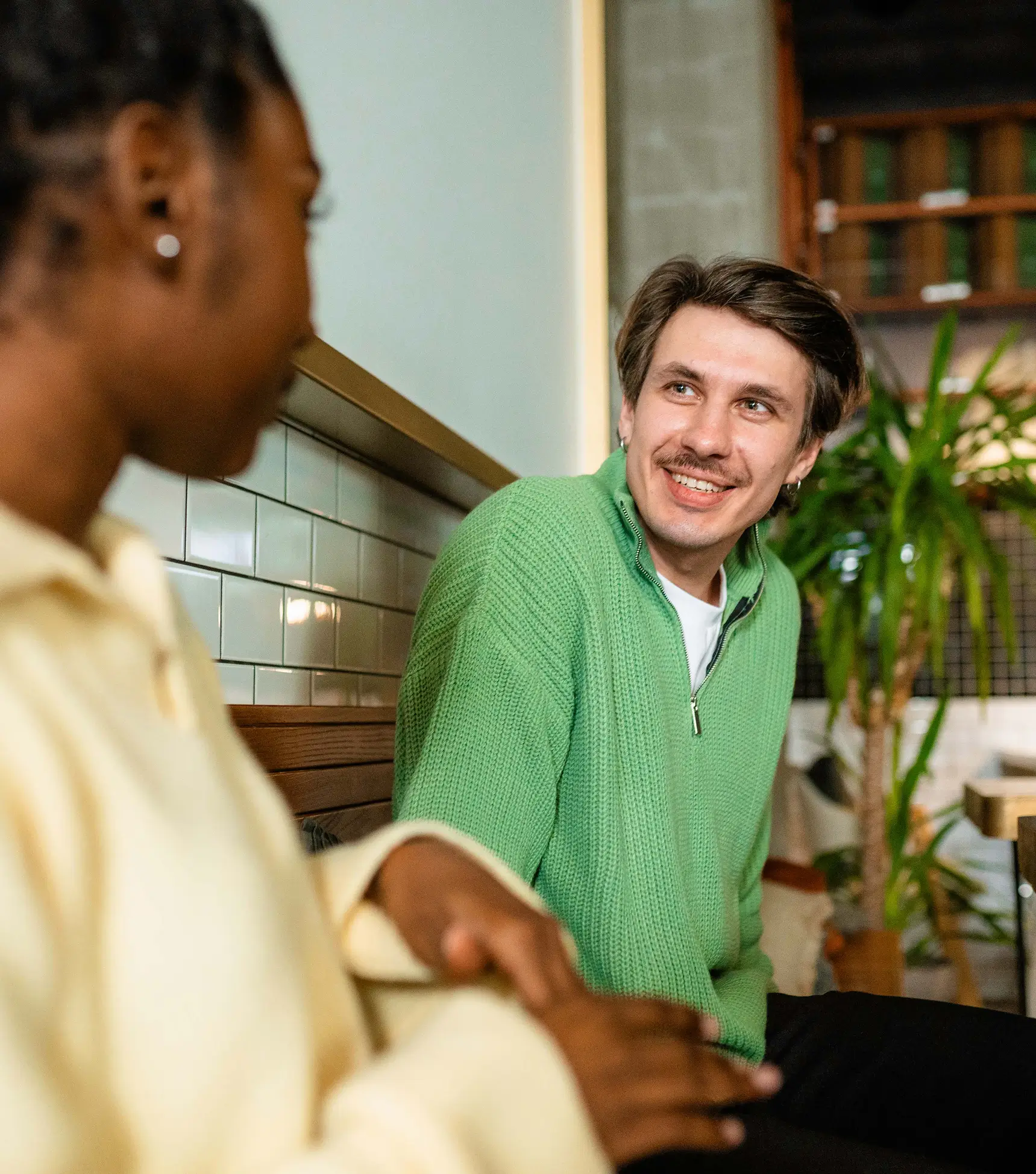 A man in a green sweater smiles and talks with a woman in a cream sweater, both sitting indoors at a New Jersey credit union near plants.