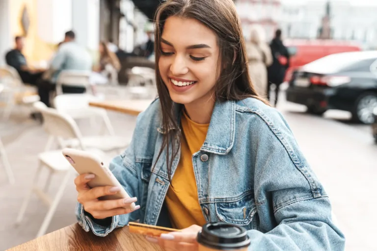 Young woman sitting at an outdoor cafe table, smiling while looking at her phone and holding a credit card, with a coffee cup in front of her.