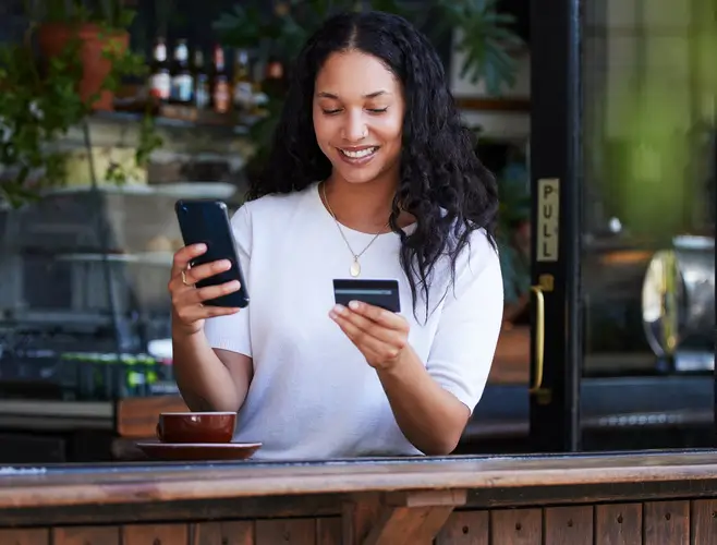 A woman stands at a counter, smiling while holding a smartphone in one hand and a credit card in the other, with a coffee cup nearby.