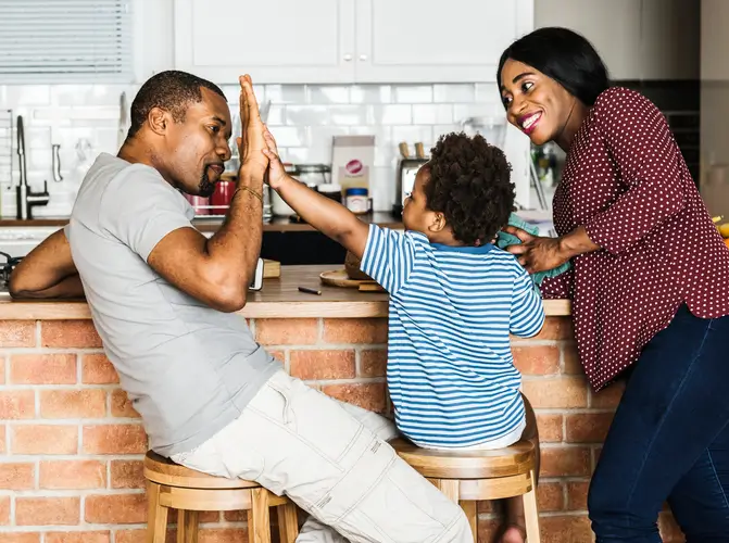 A smiling family of three in a kitchen, with a man and child giving each other a high five while a woman stands nearby, smiling at them.
