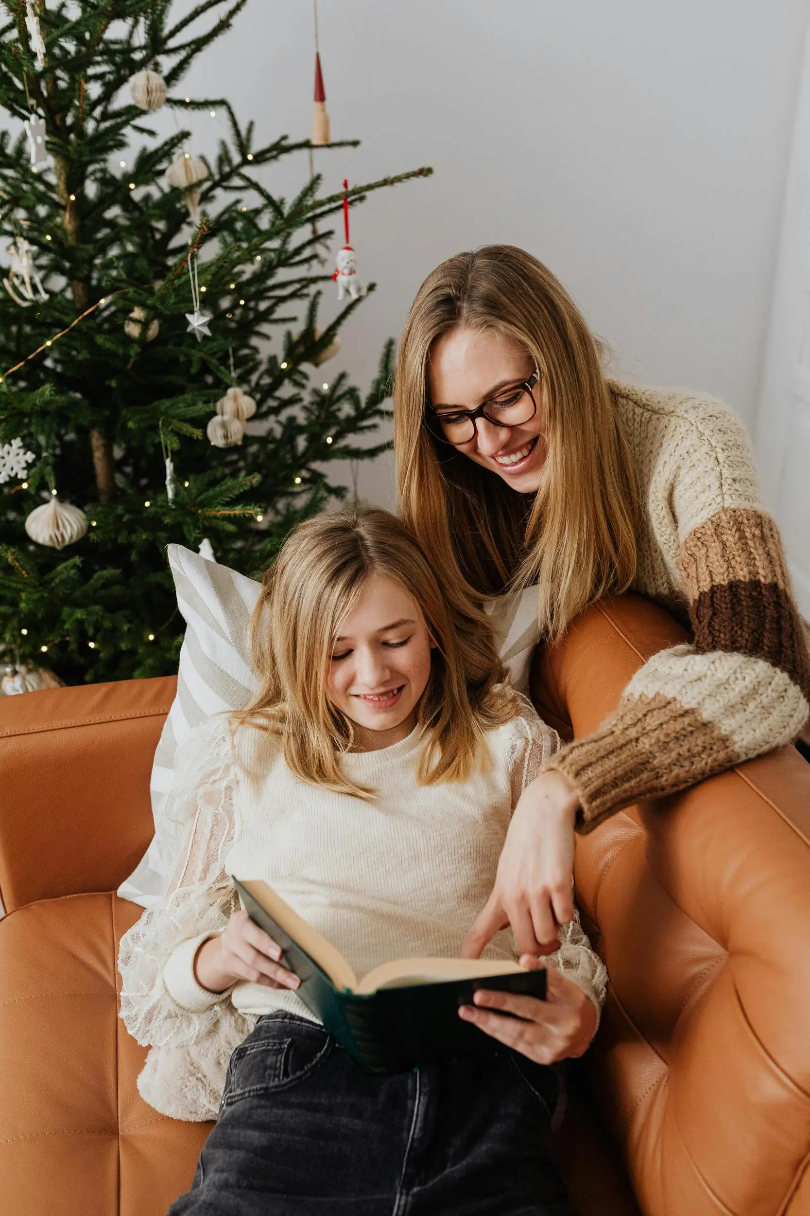 A woman and a girl sit on a brown sofa by a decorated Christmas tree, smiling as they read a book together.