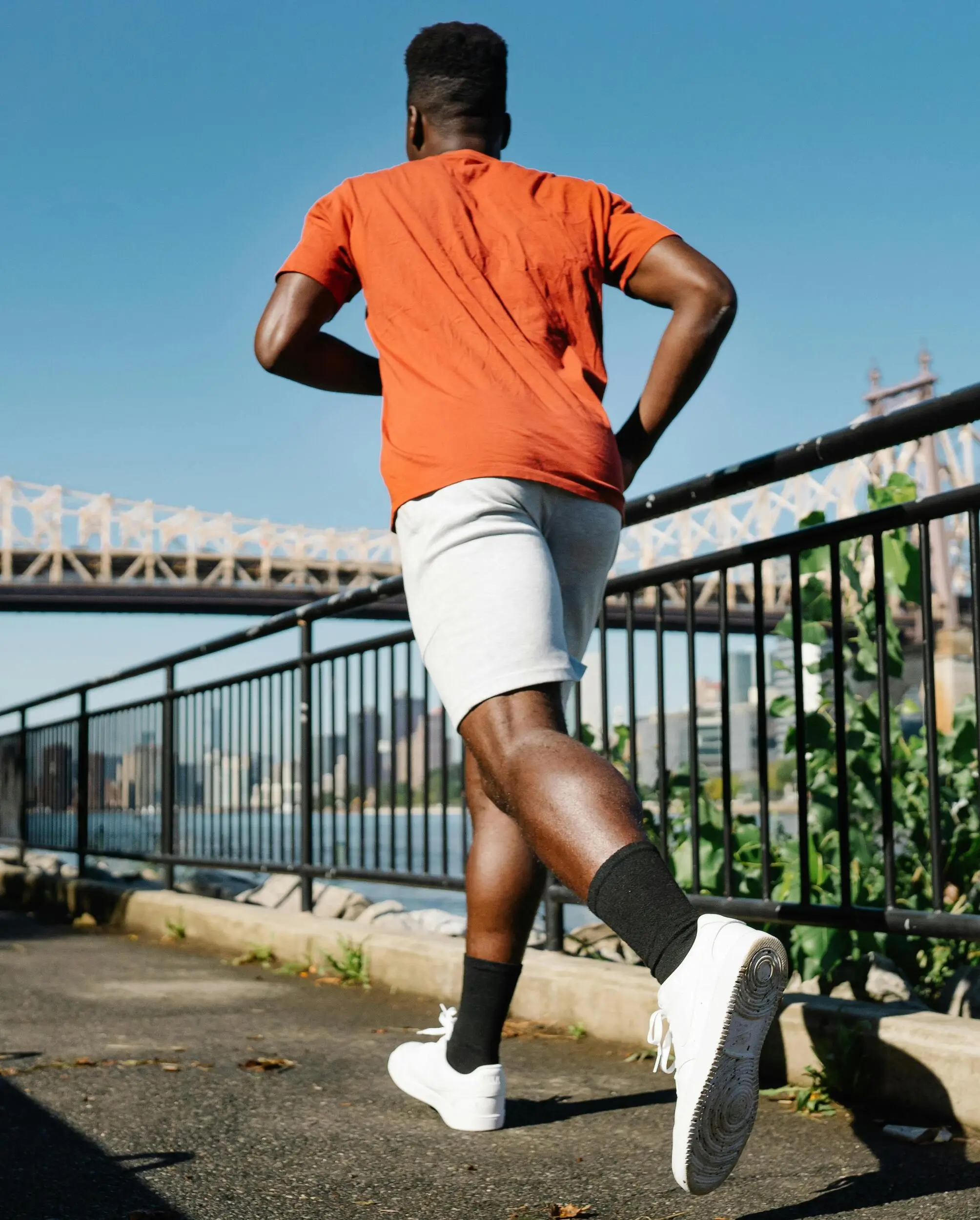 A man in an orange shirt and gray shorts jogs on a riverside path, passing by publications as a bridge and city skyline rise under a clear blue sky.
