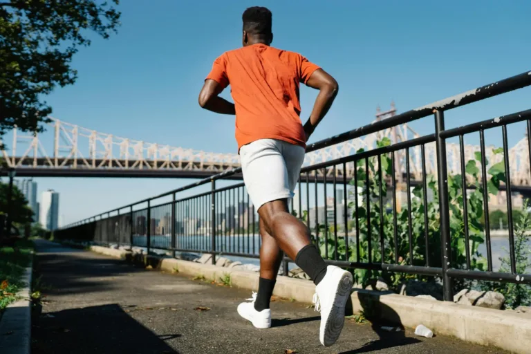 A man jogs along a riverside path with a bridge and city buildings in the background on a sunny day.