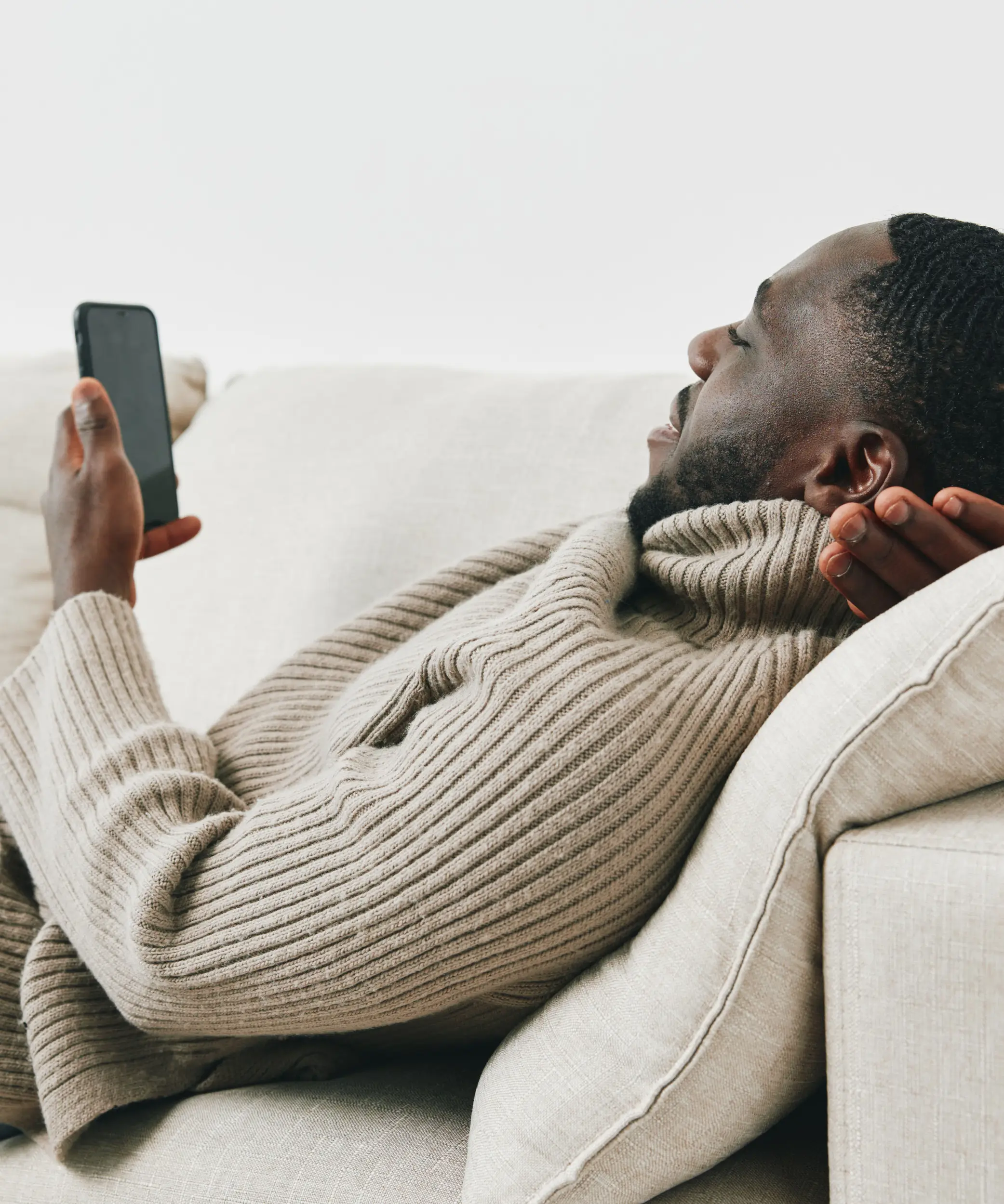 A man in a beige turtleneck sweater reclines on a light-colored sofa, smiling and looking at his smartphone.