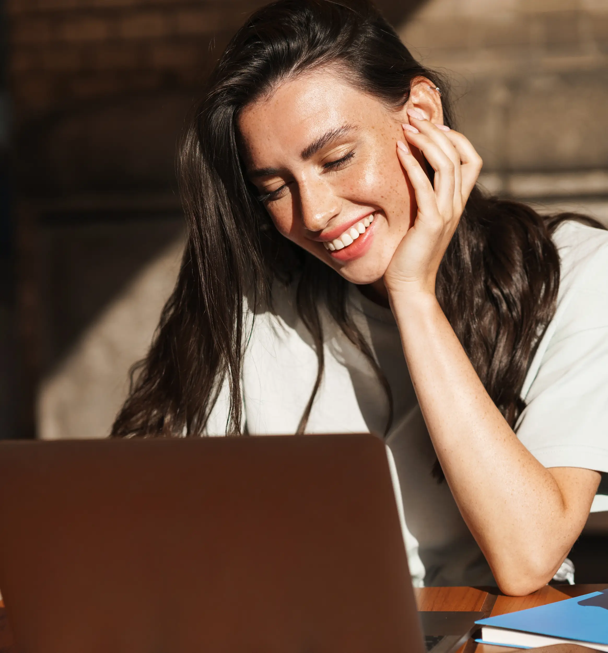 A woman with long dark hair smiles while looking at her laptop, resting her head on her hand. Sunlight shines on her face as she reviews her New York credit union account.