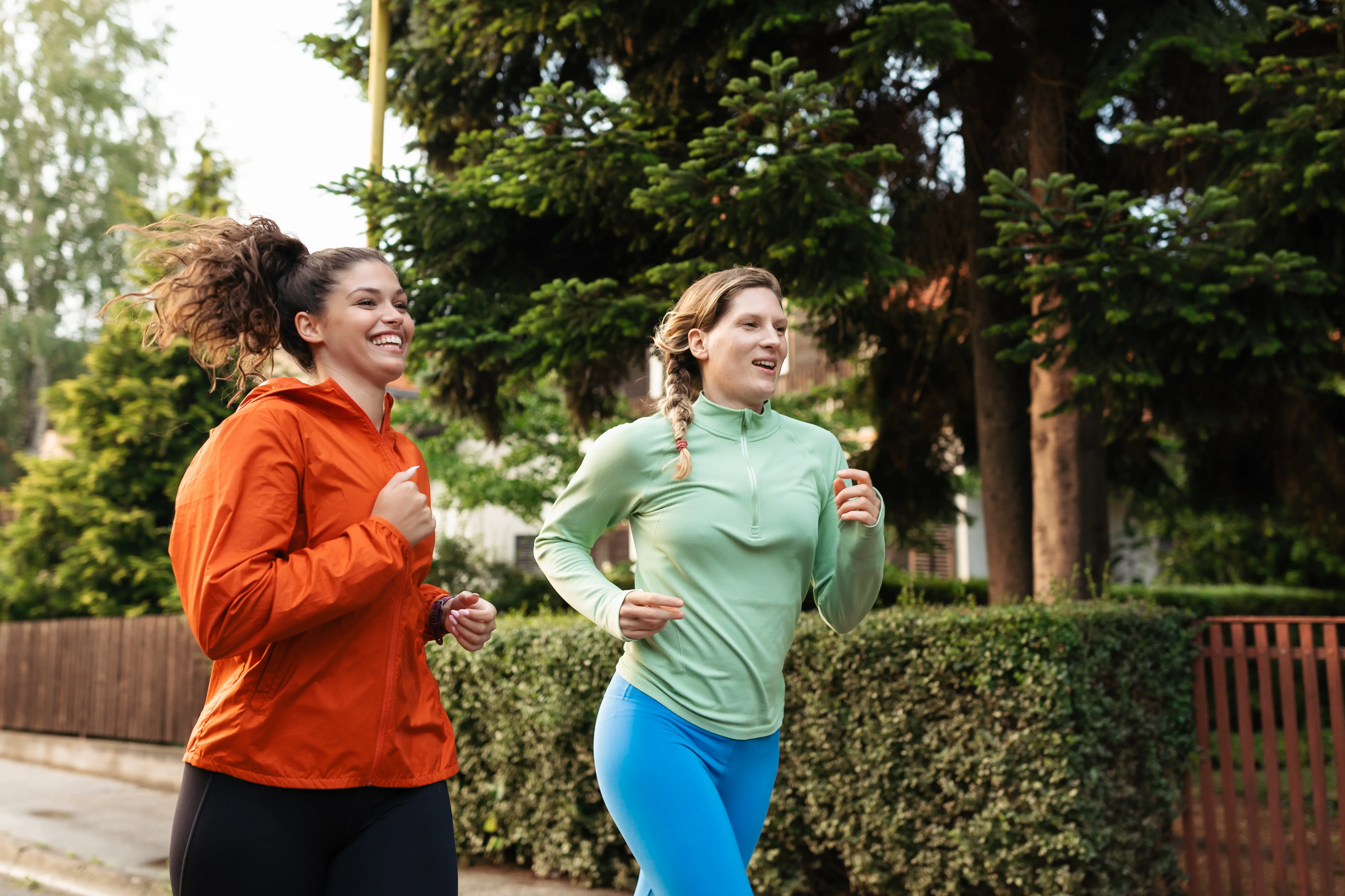 Two women in athletic clothing jogging outdoors on a sidewalk, smiling, with trees and greenery in the background—members of a New York credit union enjoying healthy lifestyles.
