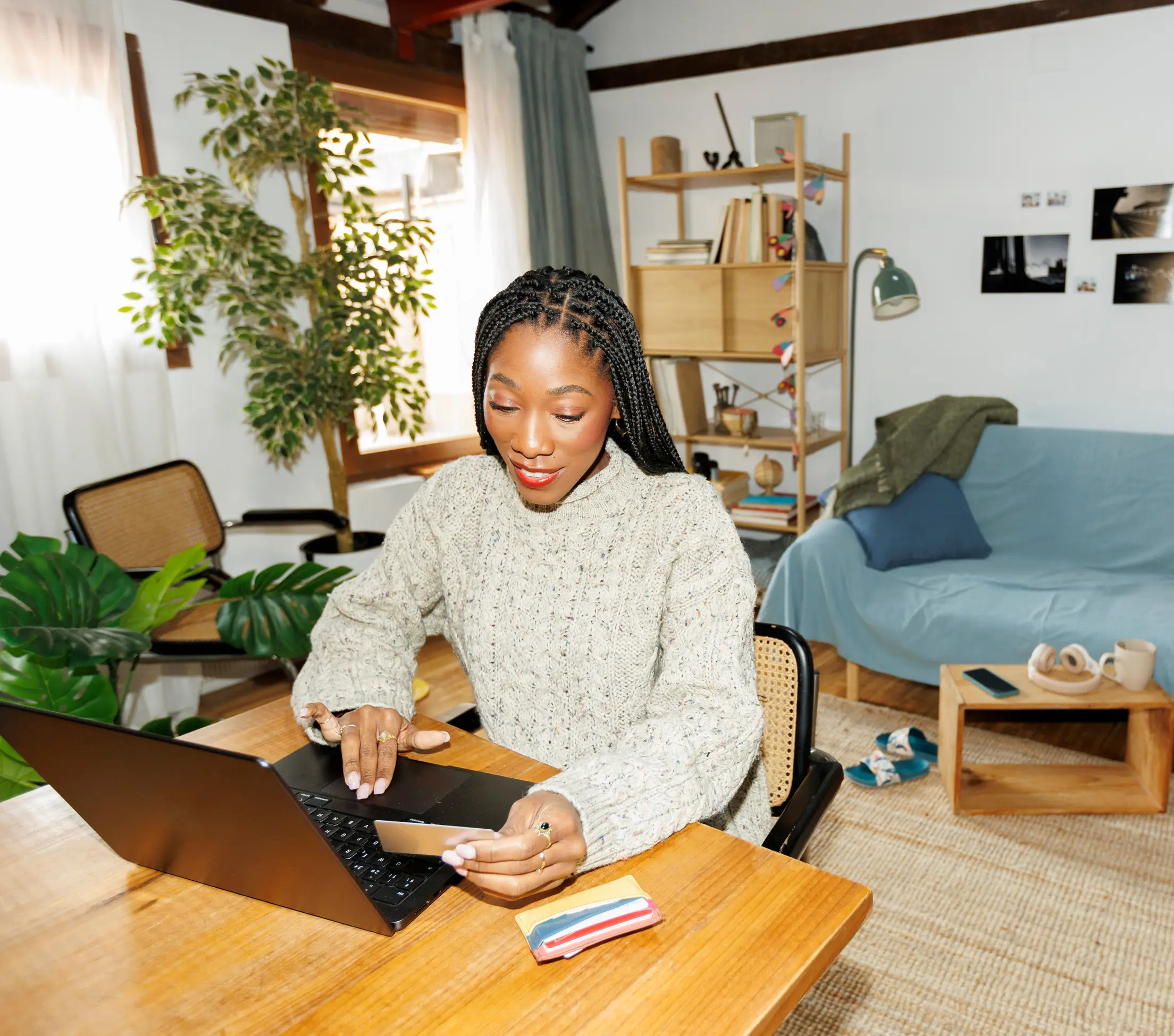 Woman with braided hair, wearing a knit sweater, sits at a desk using a laptop and holding a credit card in a cozy, well-lit room with plants and casual decor.