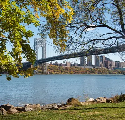 A large suspension bridge spans a wide river, with city buildings in the background—close to many New Jersey credit union branches—while trees with green leaves frame the scene.