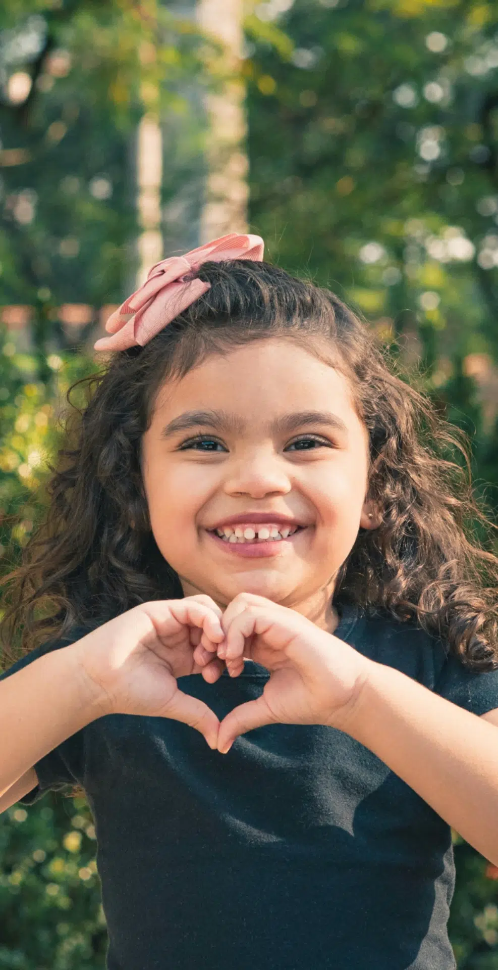 A young girl with curly hair and a pink bow, smiling and forming a heart shape with her hands outdoors, celebrates her new york credit union community in sunlight.