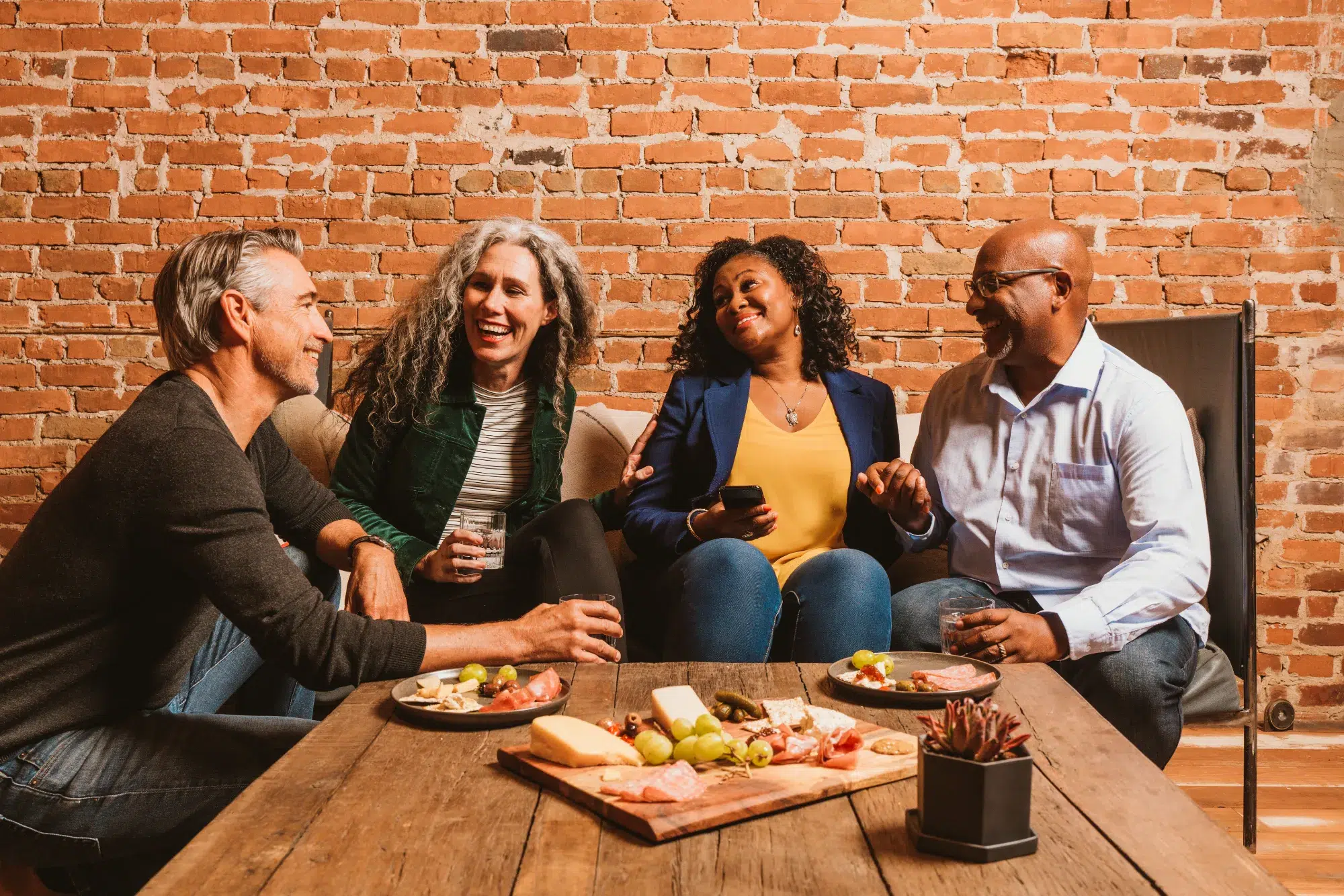 Four adults sit on a couch and chairs, smiling and laughing together, with snacks and drinks on a wooden table in front of them against a brick wall background.
