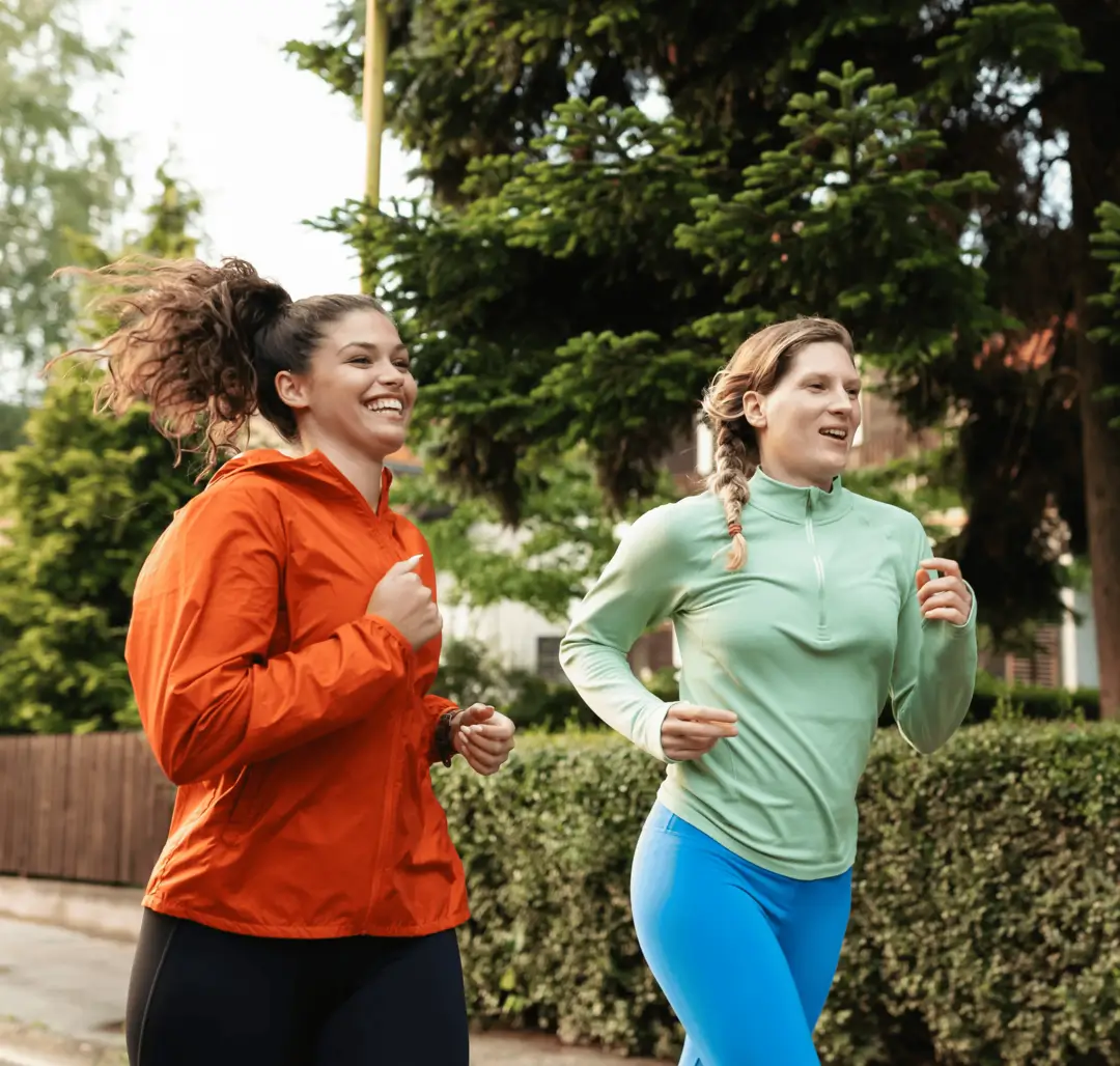 Two women jog outdoors, smiling in sporty outfits—one in an orange jacket, the other in a light green top. Trees line the path near a New Jersey credit union.