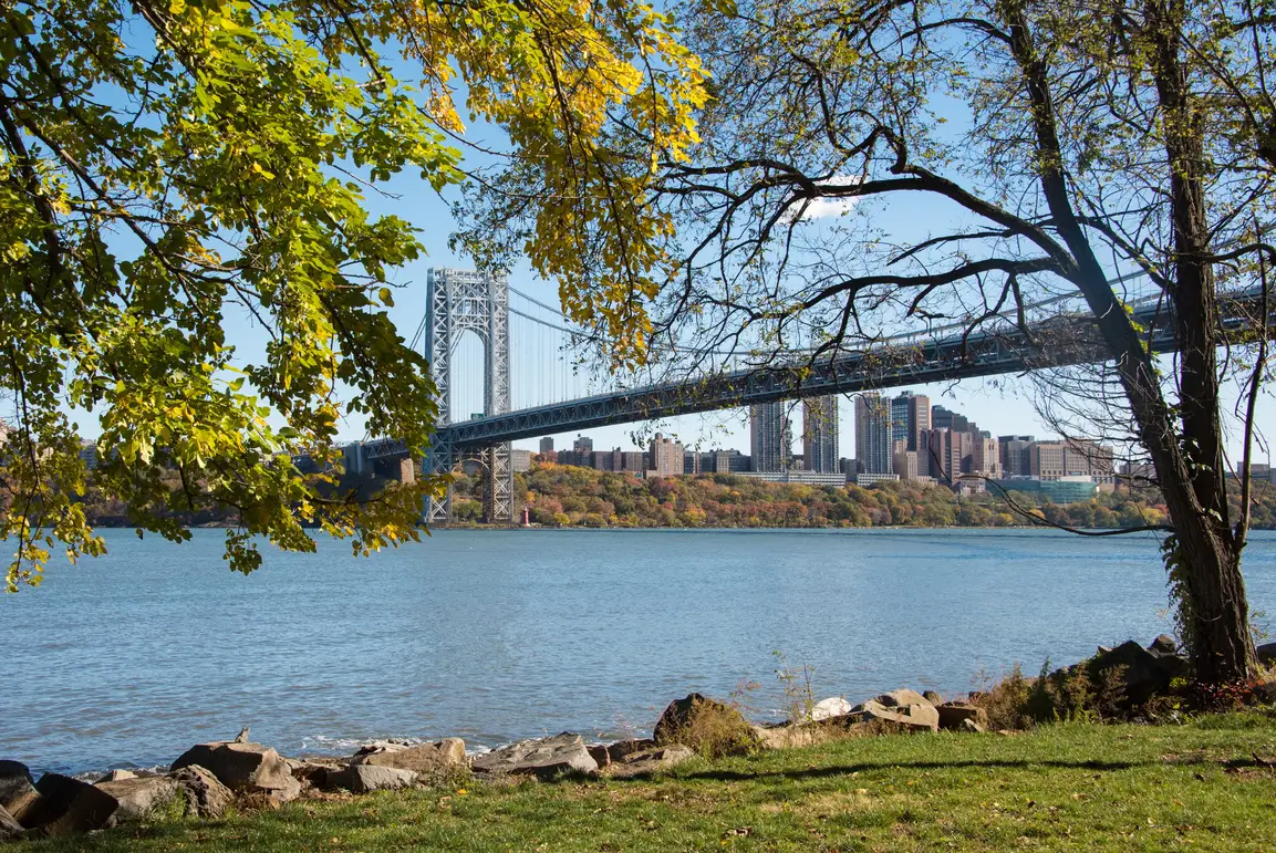 A large suspension bridge spans a wide river, with city buildings in the background—ideal for a New York or New Jersey credit union ad—framed by leafy trees on a sunny day.