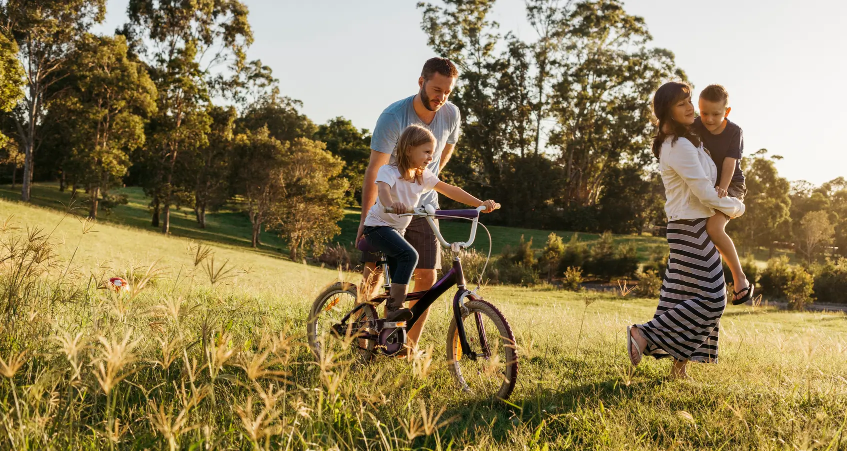A family enjoys a sunny day outdoors; in a grassy, tree-filled field, a man helps a girl ride her bike while a woman—smiling—carries their son, dreaming of joining a New York credit union.