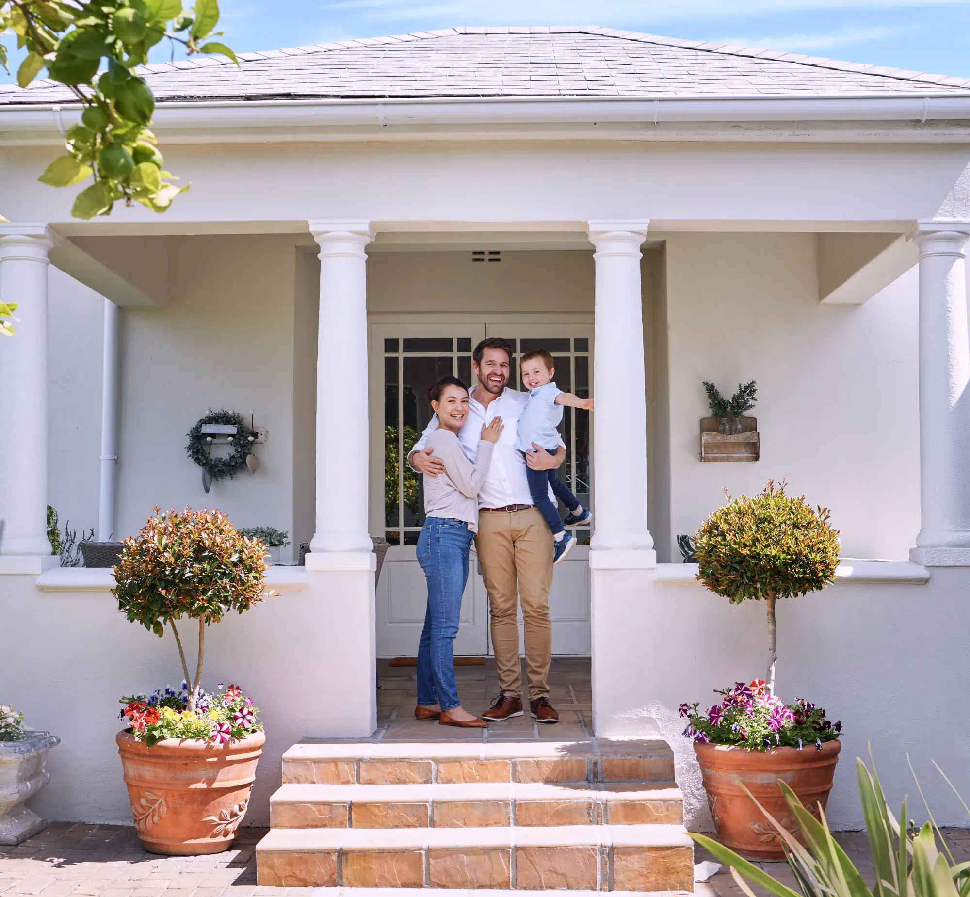 A smiling family of three stands on the front steps of a white house with columns, enjoying a sunny day—members of their trusted New Jersey credit union.