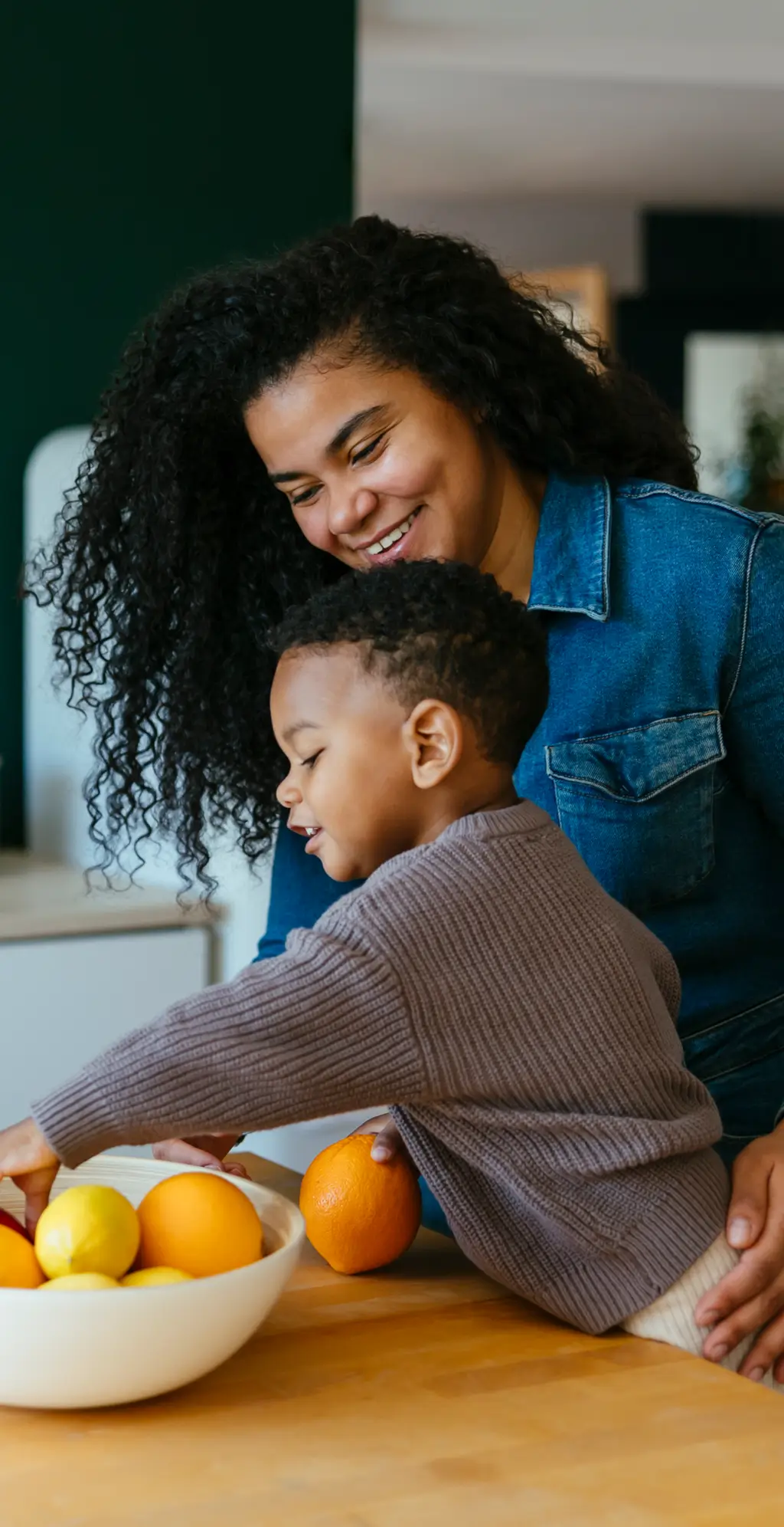 A smiling woman watches a young boy reach for oranges and lemons in a bowl on a kitchen counter, enjoying family time thanks to her New Jersey credit union.