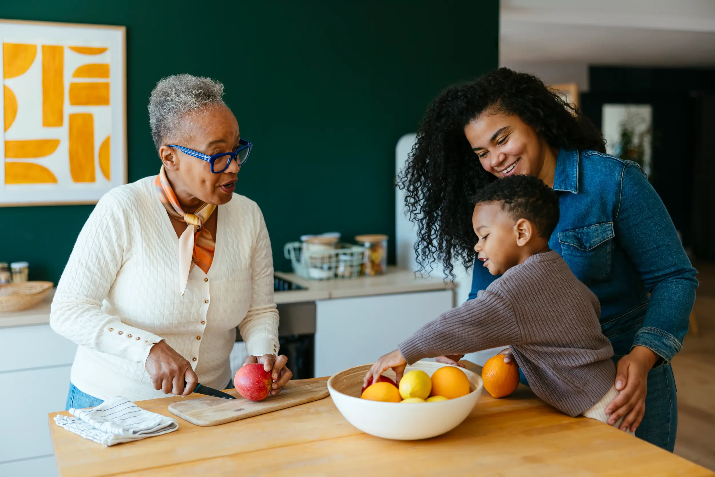 An older woman chops fruit while a young boy reaches into a bowl and a woman stands beside him, all smiling together in a modern kitchen—like family at a New York credit union event.