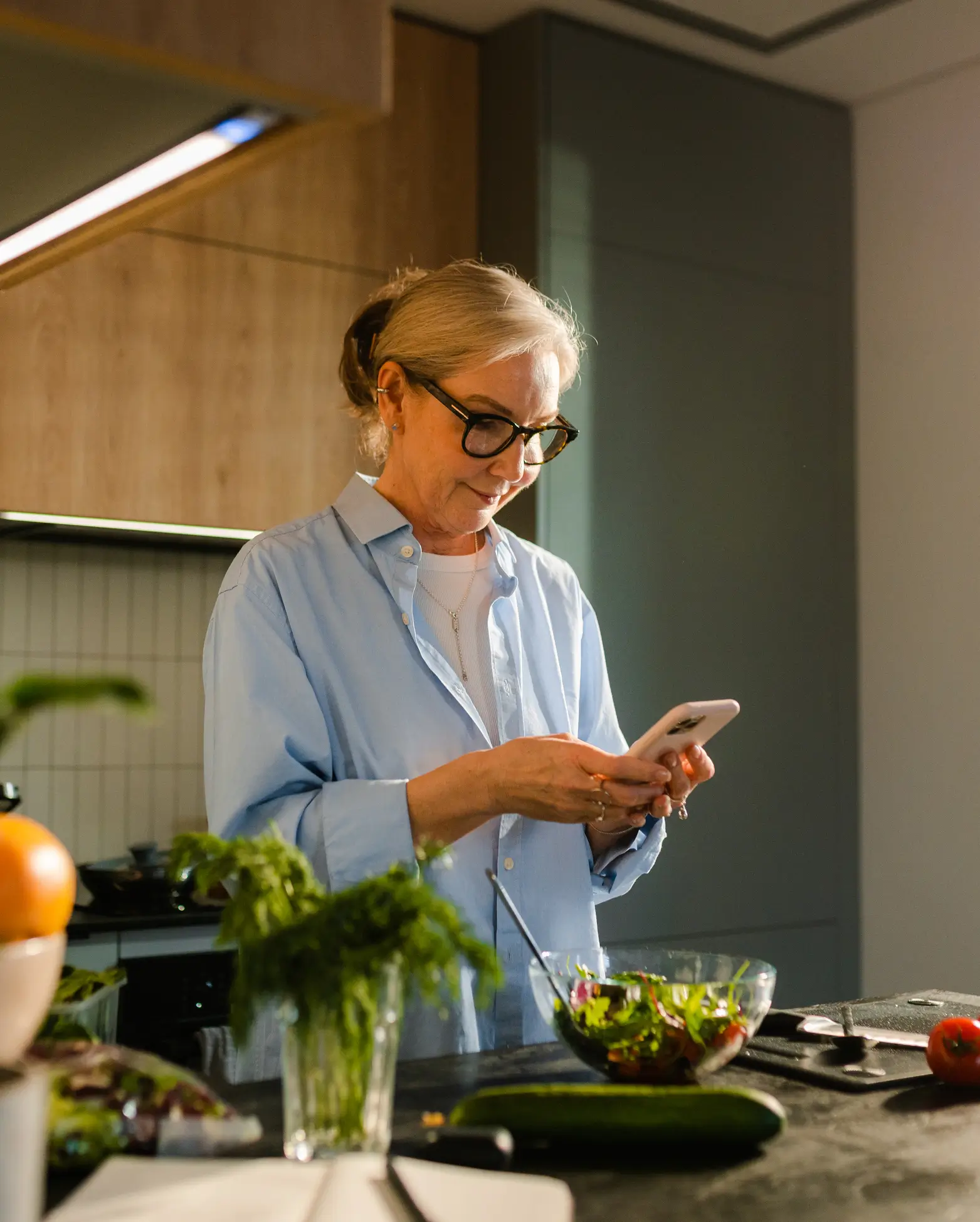 An older woman with glasses stands in a modern kitchen, checking overdraft protection on her smartphone, with fresh vegetables and herbs on the counter in front of her.