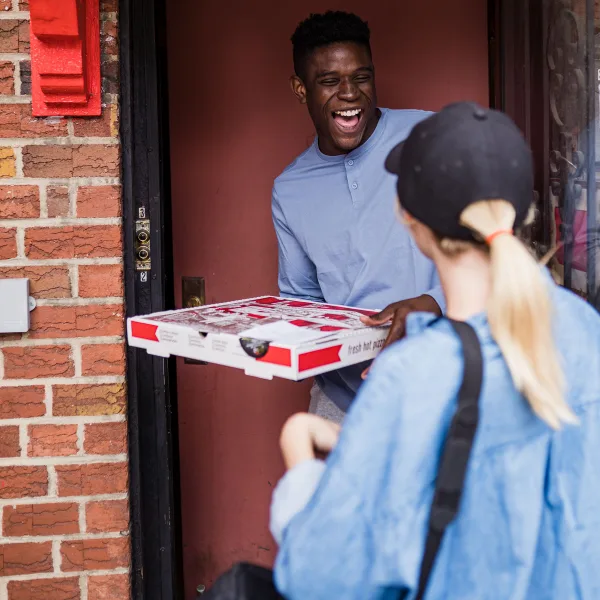 A smiling man stands at his door receiving pizza boxes from a delivery person wearing a cap and a backpack.