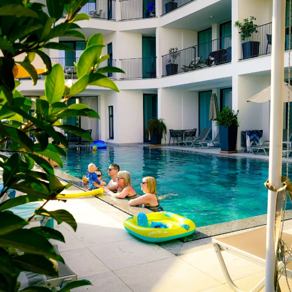 People relax in a swimming pool at a modern hotel, with balconies above and greenery in the foreground. Inflatable pool toys float nearby.