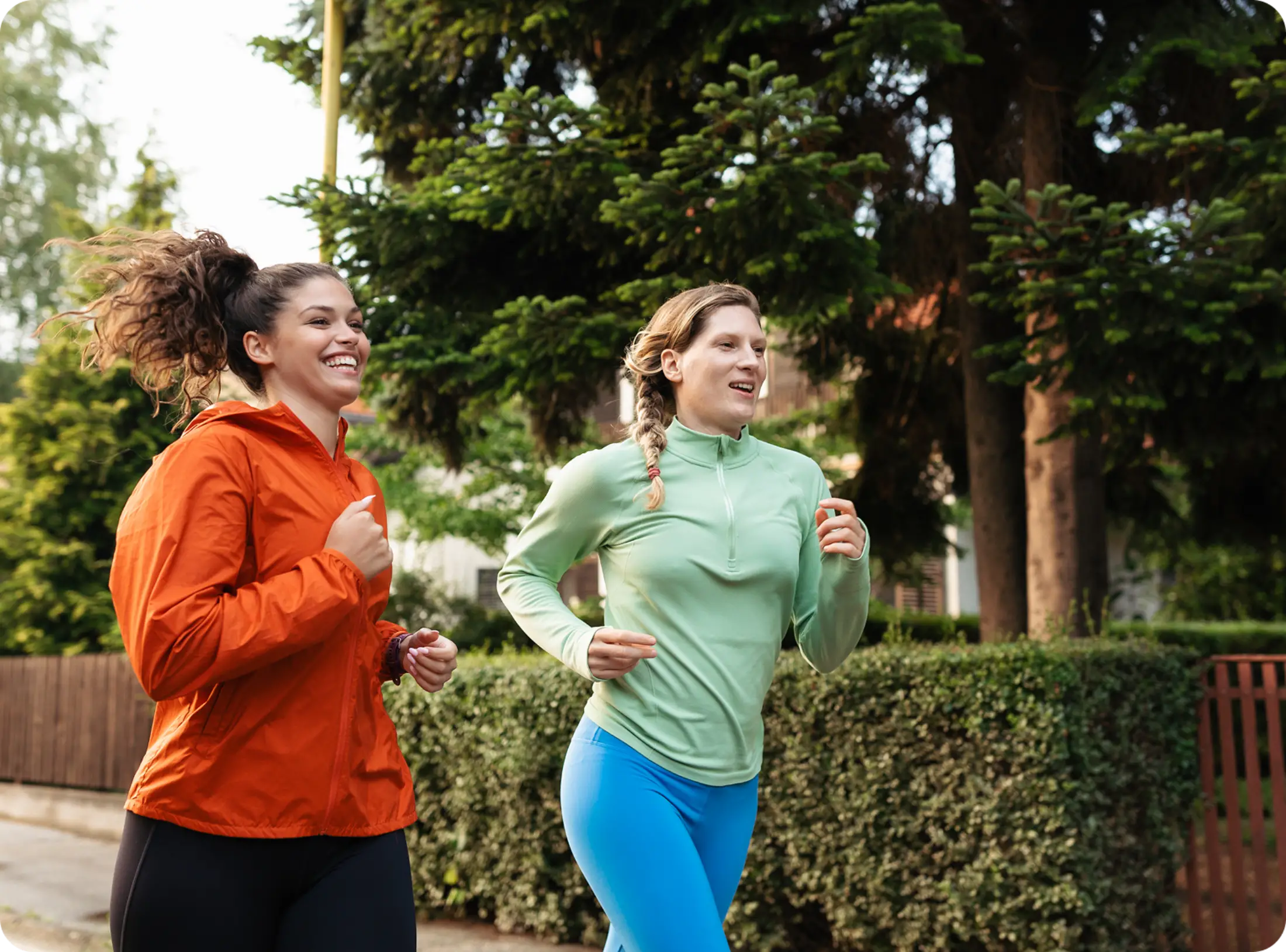Two women jog outside, both smiling. One wears an orange jacket, the other a light green top. Green trees and bushes line the background.