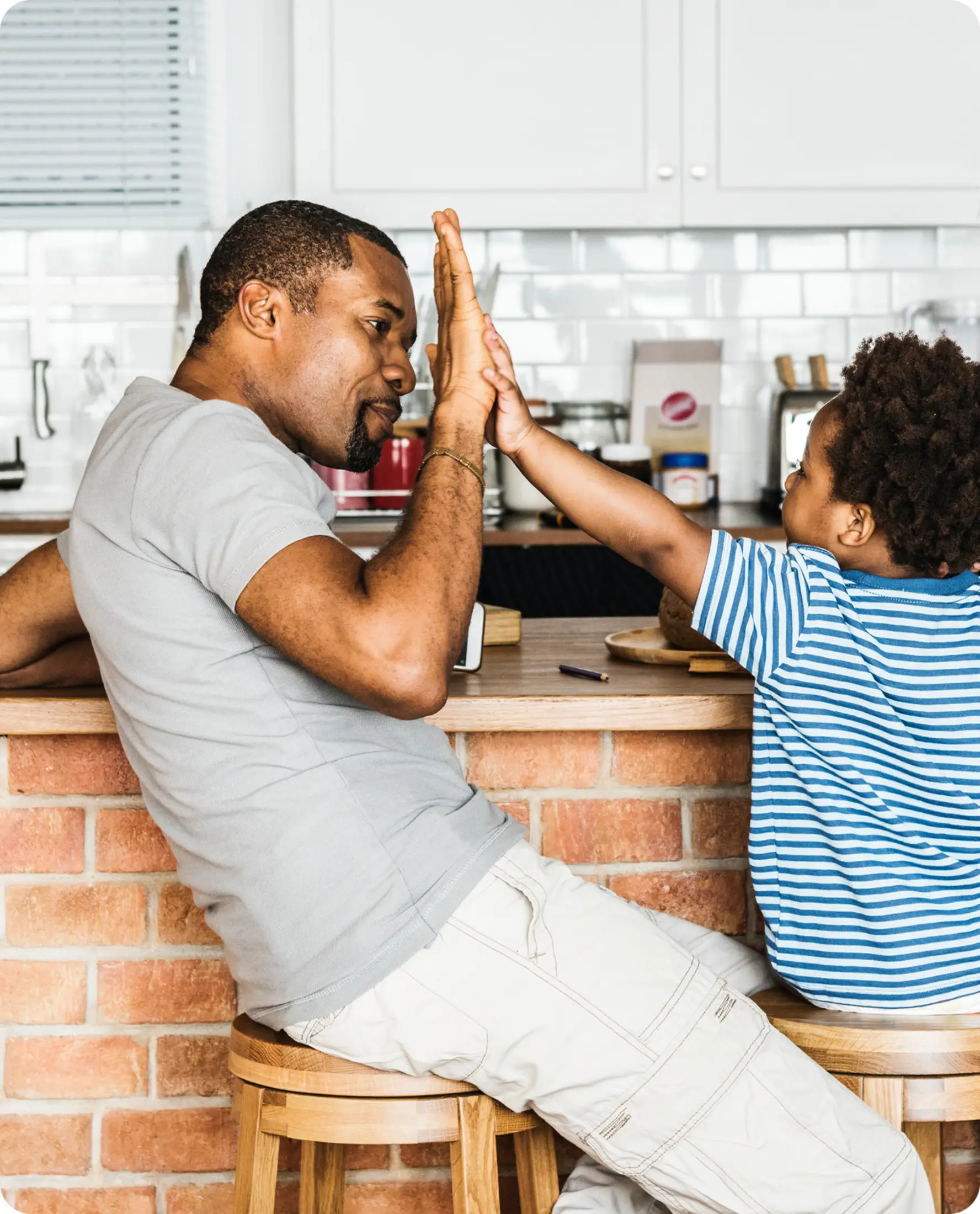 An adult and a child sit on kitchen stools, smiling and giving each other a high five over a brick counter.