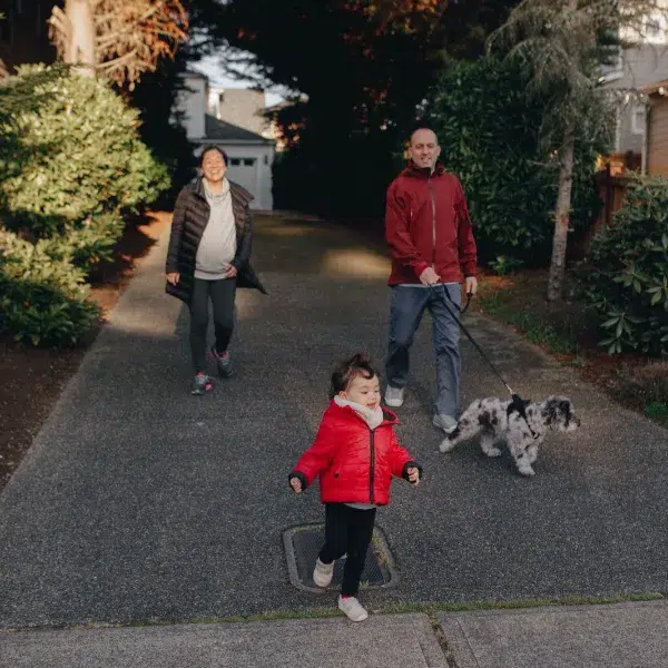 A toddler in a red jacket runs ahead on a driveway as their parents walk behind with a dog on a leash.