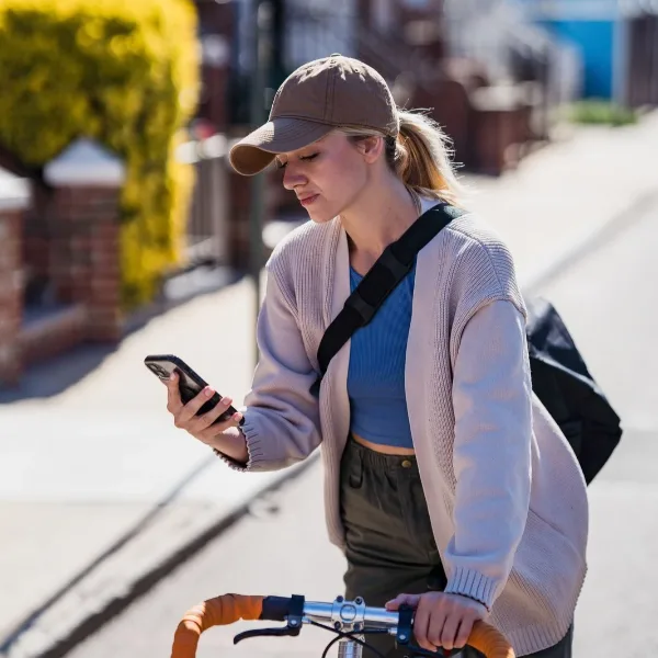 A person wearing a baseball cap and cardigan stands on a street looking at a smartphone while holding bicycle handlebars.