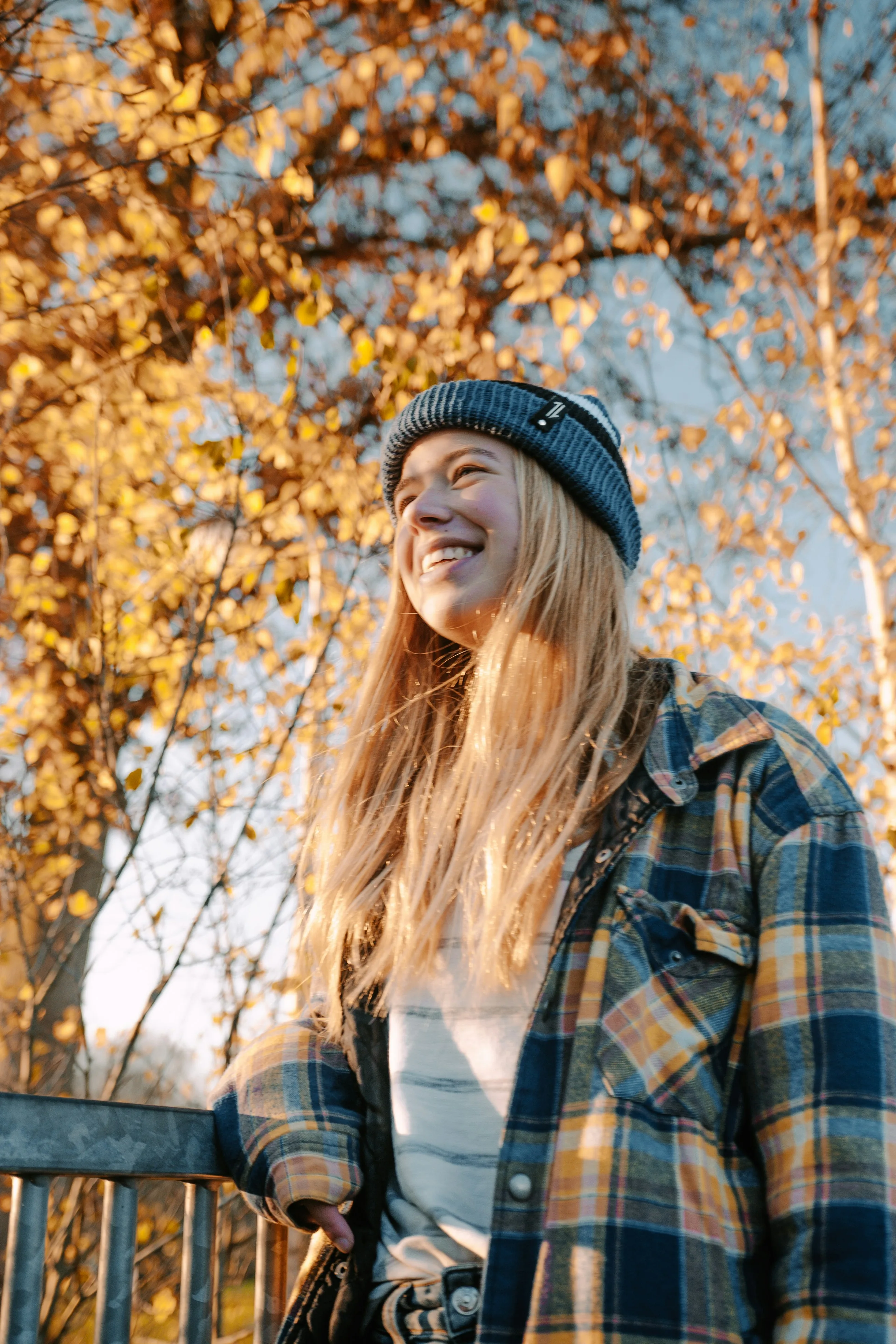 A smiling young person in a beanie and plaid shirt stands outdoors beneath autumn trees, ready to open their certificate account with their New York credit union.